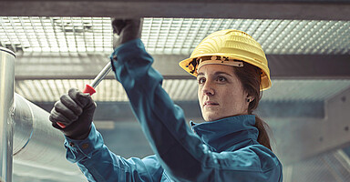 Kurzarbeit und Entgeltfortzahlung: Frau mit Schutzhelm und Schutzkleidung montiert eine Metallkonstruktion mit einem Schraubenzieher in einer Produktionshalle. Kurzarbeit und Entgeltfortzahlung: Frau mit Schutzhelm und Schutzkleidung montiert eine Metallkonstruktion mit einem Schraubenzieher in einer Produktionshalle.