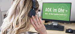 Eine Frau sitzt mit Kopfhörern vor einem Bildschirm mit der Schrift AOK im Ohr – der Arbeitgeberpodcast