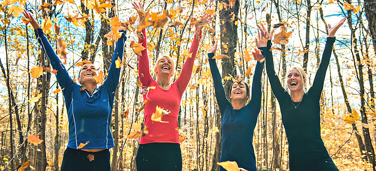 Gesund durch den Herbst: Eine Gruppe sportlich gekleideter Frauen wirft in einem Wald fröhlich Laub hoch.