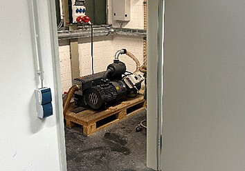 Ein Blick in einen kleinen Technikraum mit einer grauen Industriepumpe auf einer Holzpalette, aufgenommen durch einen Türrahmen.