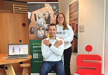 Eine AOK-Fachberaterin für Ergonomie und Bewegung führt bei einem Mitarbeiter der Sparkasse LeerWittmund den „AOK mobee-Check“ durch. Eine AOK-Fachberaterin steht hinter einem sitzenden Mitarbeiter der Sparkasse, der für eine aufrechte Haltung seine Arme vor der Brust kreuzt. Daneben ein Laptop, das Messergebnisse anzeigt.