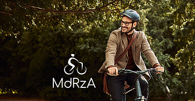 Mann mit Fahrradhelm fährt Fahrrad unter Bäumen vor einem Bürogebäude.