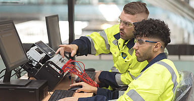 Studierende aus dem Ausland: Zwei Männer in Schutzausrüstung arbeiten am Computer. 