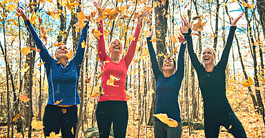 Gesund durch den Herbst: Eine Gruppe sportlich gekleideter Frauen wirft in einem Wald fröhlich Laub hoch.