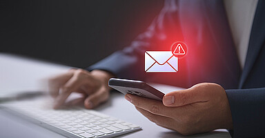 Phishing-Mails: Eine Hand, die ein Mobiltelefon in der Hand hält und darüber ist ein Briefzeichen mit einem roten Ausrufezeichen