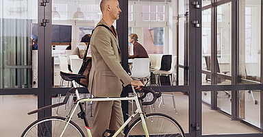 Grenzmanagement: Mann im Anzug schiebt ein Fahrrad durch ein modernes Büro mit Glaswänden.