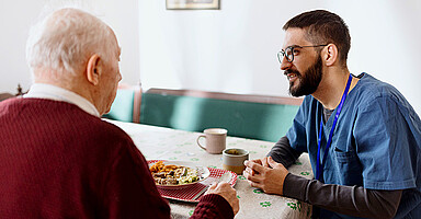 Ausländische Fachkräfte: Ein Pfleger sitzt bei einem Senior, der gerade isst