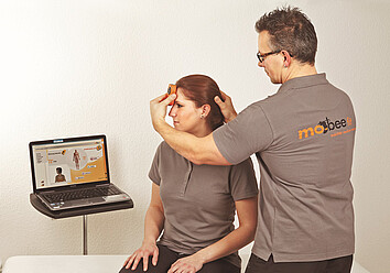 Messvorgang beim „AOK mobee-Check“ Ein AOK-Mitarbeiter hält ein kleines Messgerät an die Stirn einer jungen Frau. Beide schauen auf ein Laptop, das die Messergebnisse anzeigt.