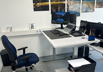 Ein ergonomischer Schreibtisch mit einem Bürostuhl. Auf dem Schreibtisch stehen ein Laptop, zwei Monitore und ein Telefon. An der Wand hängen mehrere gerahmte Bilder.