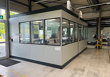 Ein freistehendes, modernes Bürogebäude mit großen Fenstern in einer Fabrikhalle.
