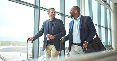 Entsendungen in Abkommensstaaten 2026: Zwei Reisende unterwegs zum Gate im Flughafen. Entsendungen in Abkommensstaaten 2026: Zwei Reisende unterwegs zum Gate im Flughafen.