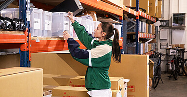 Arbeitszeiterfassung ist Pflicht: Frau greift nach Kiste in einem Regallager.