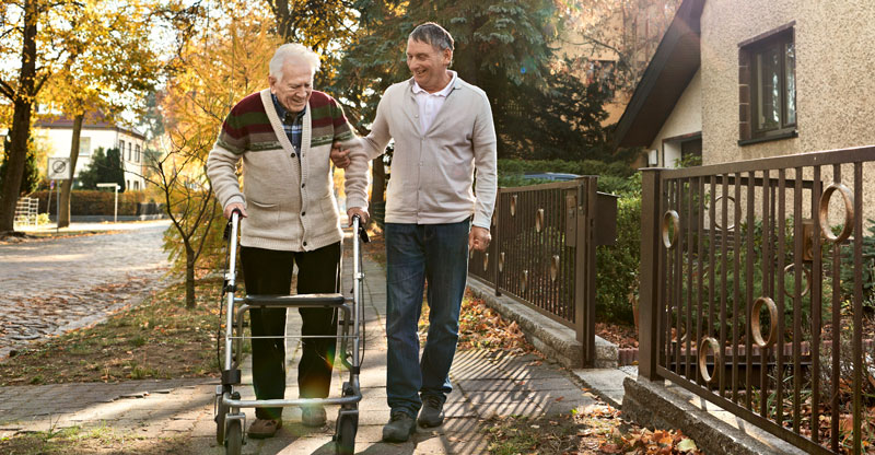 Ein Mann geht mit einer pflegebedürftigen Person am Rollator spazieren.