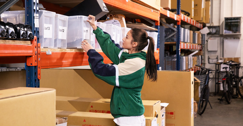 Arbeitszeiterfassung ist Pflicht: Frau greift nach Kiste in einem Regallager. Arbeitszeiterfassung ist Pflicht: Frau greift nach Kiste in einem Regallager.