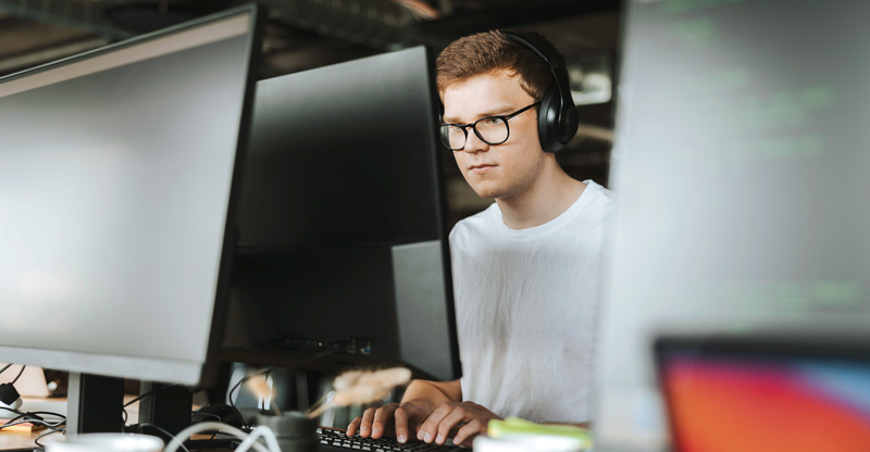 Neurodiversität: Blick auf einen jungen Mann durch Monitore hindurch, der hochkonzentriert ist und Kopfhörer trägt.