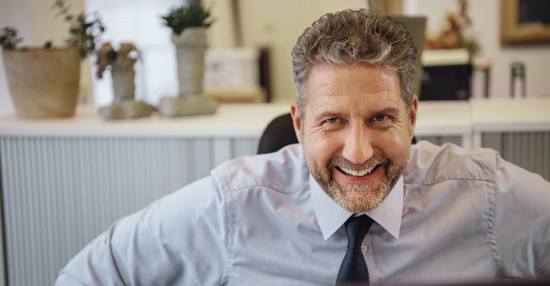 Resilienz im Arbeitsalltag: Lächelnder Mann im Büro sitzt am Schreibtisch und schaut in die Kamera