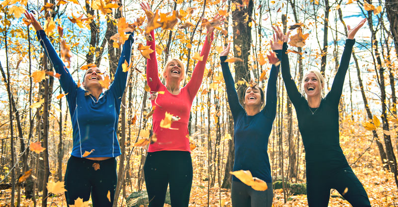 Gesund durch den Herbst: Eine Gruppe sportlich gekleideter Frauen wirft in einem Wald fröhlich Laub hoch.
