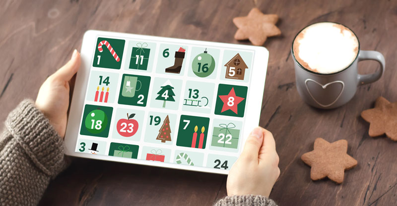 Eine Person hält ein Tablet, das einen digitalen Adventskalender anzeigt. Daneben sind eine Tasse Kaffee und Plätzchen auf einem Holztisch zu sehen.