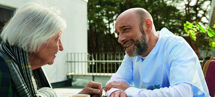 Die Mehrzahl der Pflegekräfte arbeitet in Teilzeit. Eine ältere Dame und ein Pfleger sitzen am Tisch und unterhalten sich.