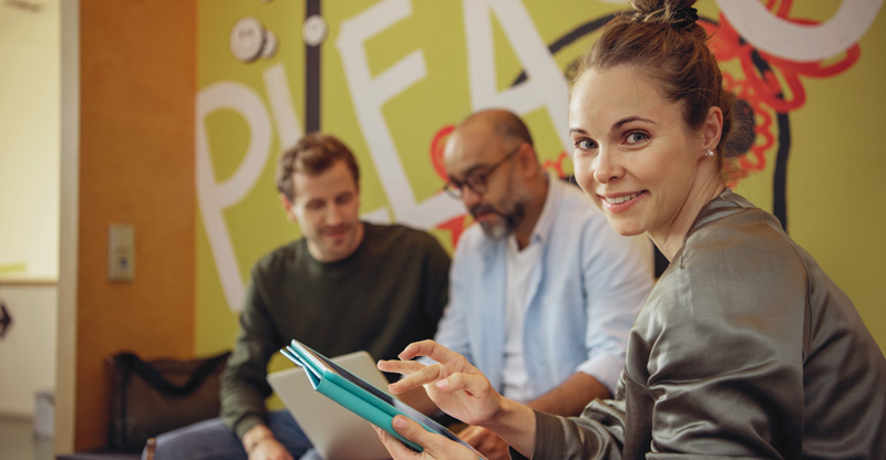 Zu sehen sind zwei Männern und eine Frau. Die Frau hält ein Tablet in der Hand und lächelt.
