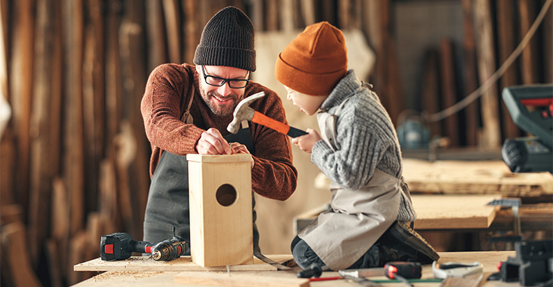 Zu sehen sind ein Mann und ein kleines Kind in einer Werkstatt, die ein Vogelhaus bauen.