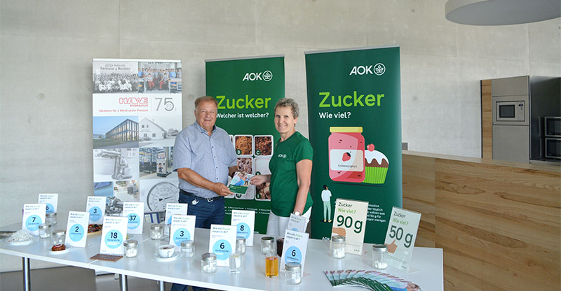 Zwei Personen stehen an einem Ausstellungstisch, der Zuckergehalt in verschiedenen Lebensmitteln (dargestellt durch Zuckerwürfel in Gläsern) zeigt. Im Hintergrund sind AOK-Banner zu sehen, eines fragt: "Zucker Wie viel?".