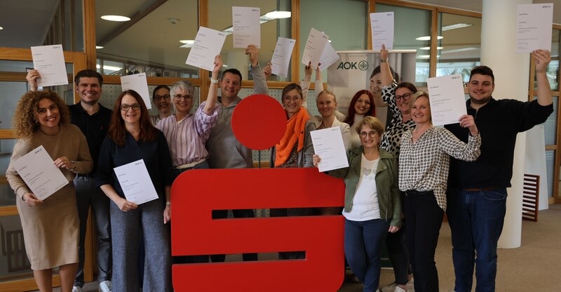 Gruppenfoto: Mitarbeitende des Sparkassenverbands Bayern halten ihre AOK-Zertifikate hoch, in der Mitte ein großes rotes Sparkassen-Logo.
