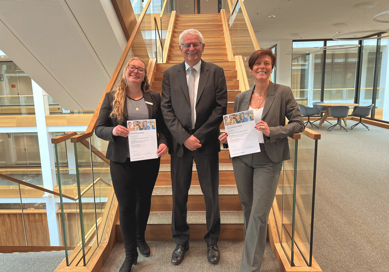 Lilli Mazurek, Frank Fastenau und Kerstin Akkermann (von links) freuen sich über die Qualifikation zu „Betrieblichen Gesundheitscoaches“. Links und rechts von einem lächelnden Mann stehen zwei lächelnde Frauen, die Zertifikate in die Kamera halten.