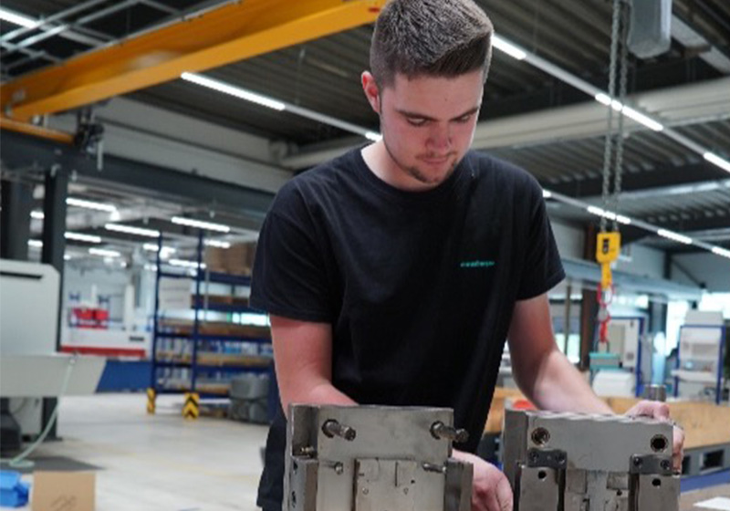 Ein junger Mann in einem schwarzen T-Shirt mit kurzem Haar, der in einer Werkstatt zwei Metallteile bearbeitet.