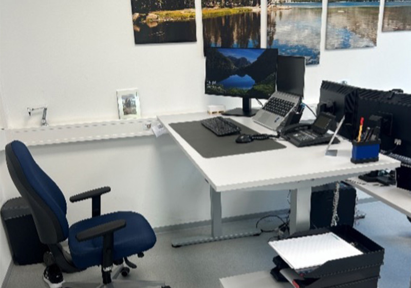 Ein ergonomischer Schreibtisch mit einem Bürostuhl. Auf dem Schreibtisch stehen ein Laptop, zwei Monitore und ein Telefon. An der Wand hängen mehrere gerahmte Bilder.