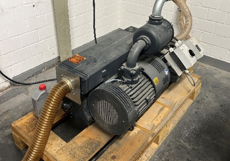 Eine große, graue Industriepumpe mit Schläuchen steht auf einer Holzpalette in einem Lagerraum.
