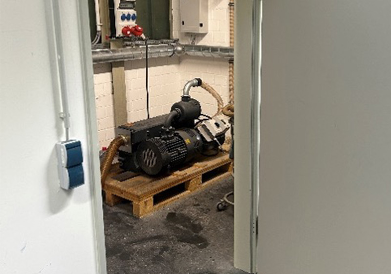 Ein Blick in einen kleinen Technikraum mit einer grauen Industriepumpe auf einer Holzpalette, aufgenommen durch einen Türrahmen.