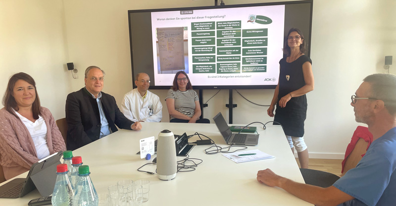 BDH-Klinik Vallendar: Eine Gruppe aus drei Frauen und drei Männern in einer Workshopsituation sitzen an einem Tisch vor einem großen Bildschirm.