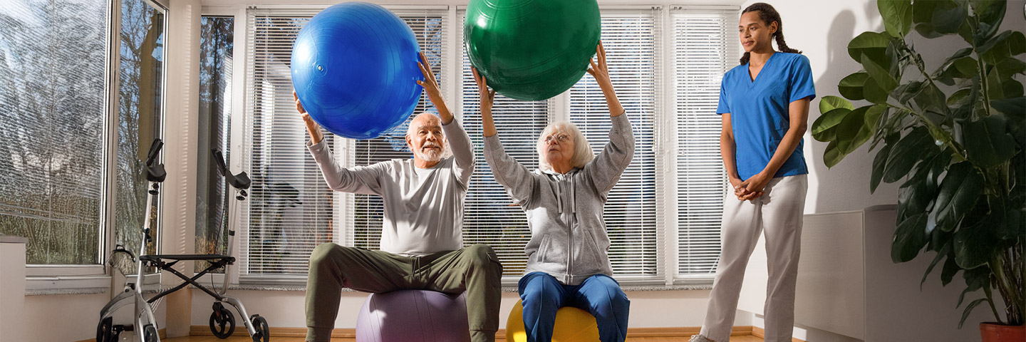Gesundheit in der Altenpflege fördern: Zwei ältere Personen führen Übungen mit Gymnastikbällen aus und werden von einer weiblichen Pflegekraft dabei beobachtet.