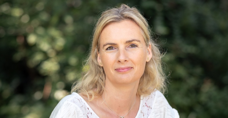 Simone Bücking Simone Bücking sitzt lächelnd auf einer Holzbank im Grünen und trägt eine weiße Bluse mit Spitzendetails