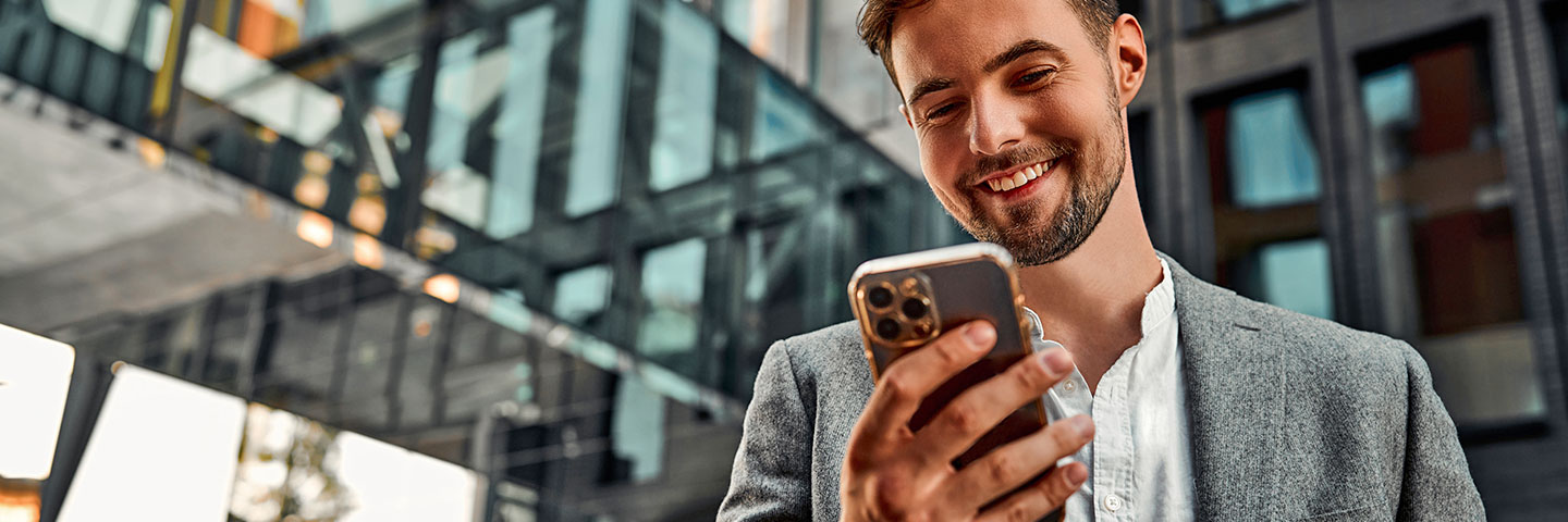 Ein Mann steht vor einem Gebäude und schaut freudig auf sein Mobiltelefon.