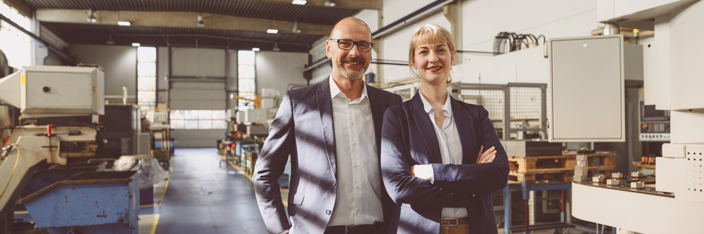 Mann und Frau in Businesskleidung stehen in einer Fabrikhalle.