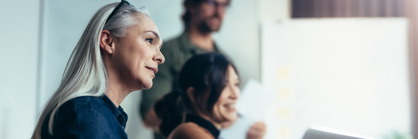 Eine mittelalte Frau mit langen, grauen Haaren sitzt lächelnd an einem Konferenztisch und blickt interessiert zur Seite, während sie an einem Meeting teilnimmt.