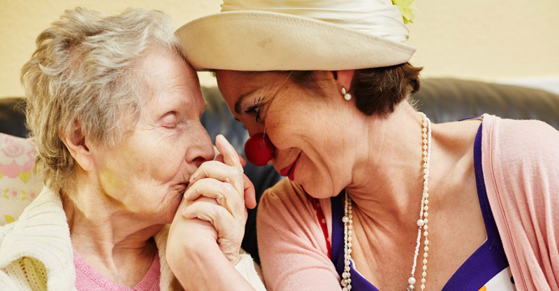 Humor in der Pflege: Ältere Dame küsst Hand von Frau mit Clownsnase