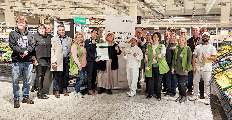 Goldzertifizierung Zu sehen ist eine Gruppe aus Mitarbeitenden in einem Supermarkt. Eine Frau aus der Gruppe hält ein Zertifikat in der Hand. Kooperation mit AOK führte zum vierten Mal zur Goldzertifizierung.