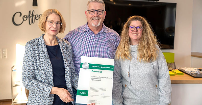 Zertifizierung als gesundes Unternehmen in Silber Zu sehen sind zwei Frauen und ein Mann die das Zertifikat in der Hand halten.
