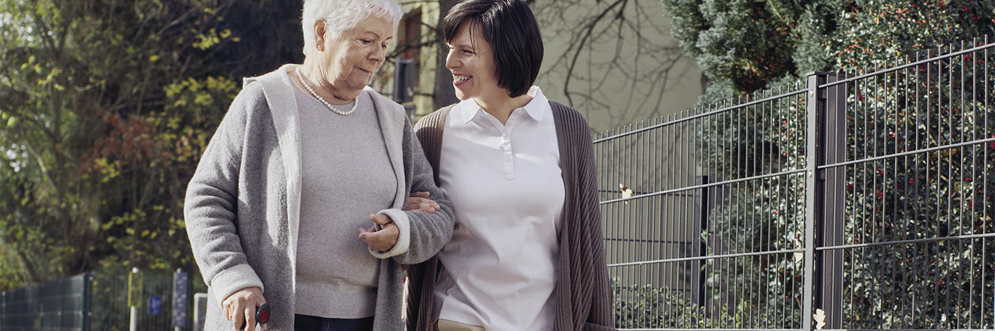 Eine Pflegekraft begleitet eine pflegebedürftige Frau auf einem Fußweg.