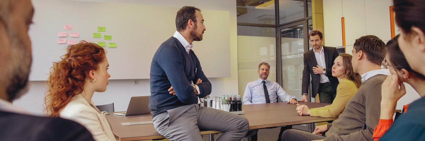 Meetingsituation mit sitzenden und stehenden Teilnehmenden in einem Besprechungsraum.