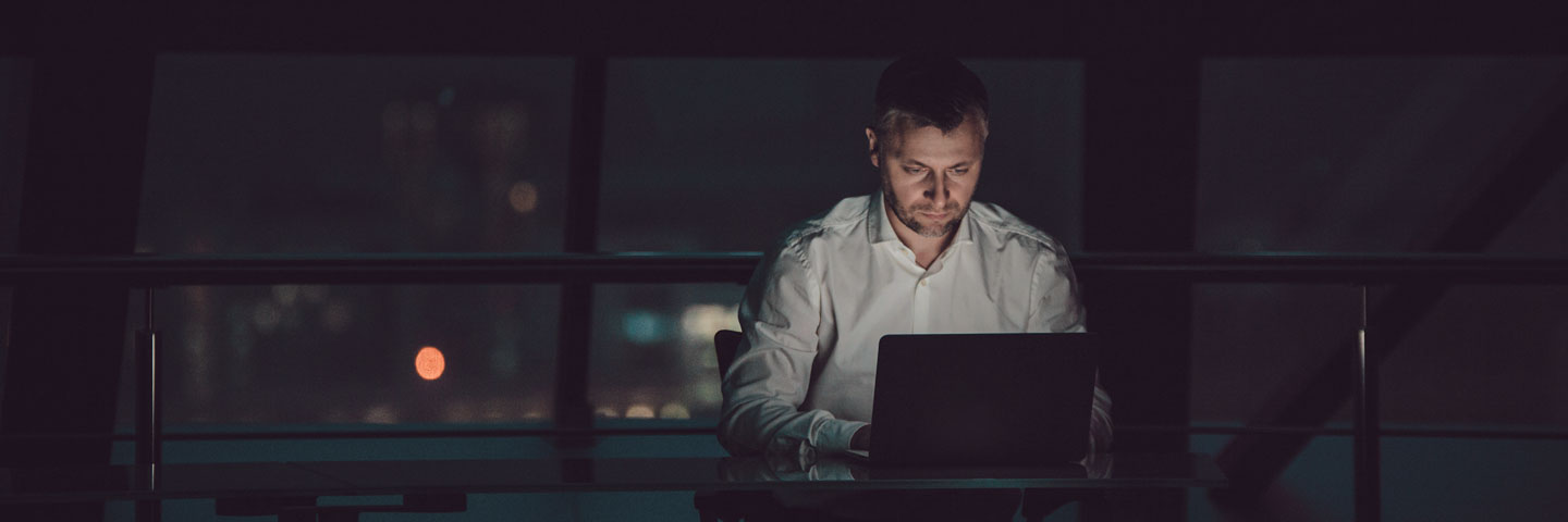 Mann sitzt nachts im Büro im Dunkeln, nur beleuchtet vom Schein des Monitors. 