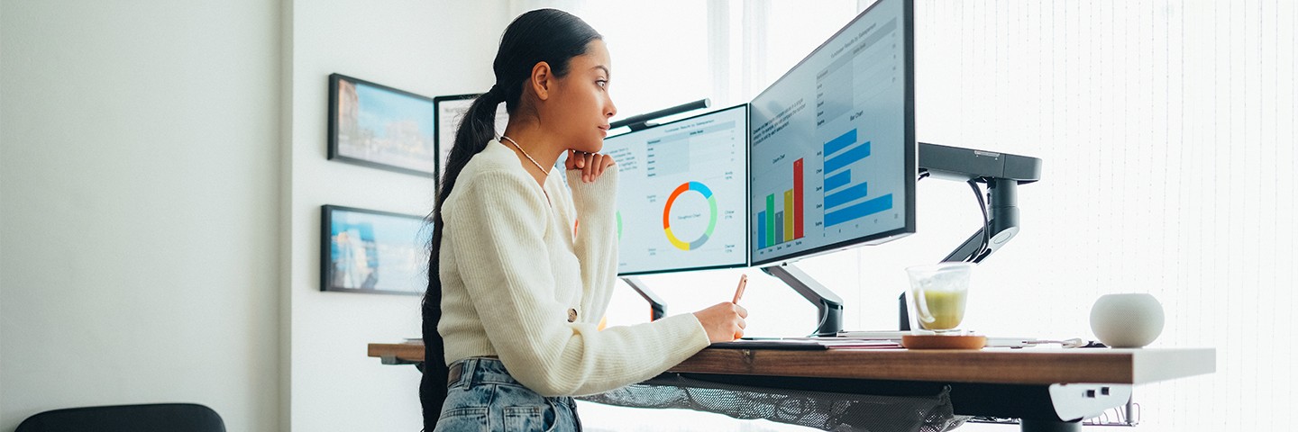 Ergonomie am Arbeitsplatz: eine Frau blickt stehend auf zwei Monitore an einem höhenverstellbaren Schreibtisch Ergonomie am Arbeitsplatz: eine Frau blickt stehend auf zwei Monitore an einem höhenverstellbaren Schreibtisch