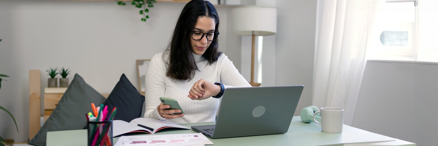Selbstorganisation im Homeoffice: Junge Frau am heimischen Schreibtisch checkt Handy und Uhrzeit. Selbstorganisation im Homeoffice: Junge Frau am heimischen Schreibtisch checkt Handy und Uhrzeit.