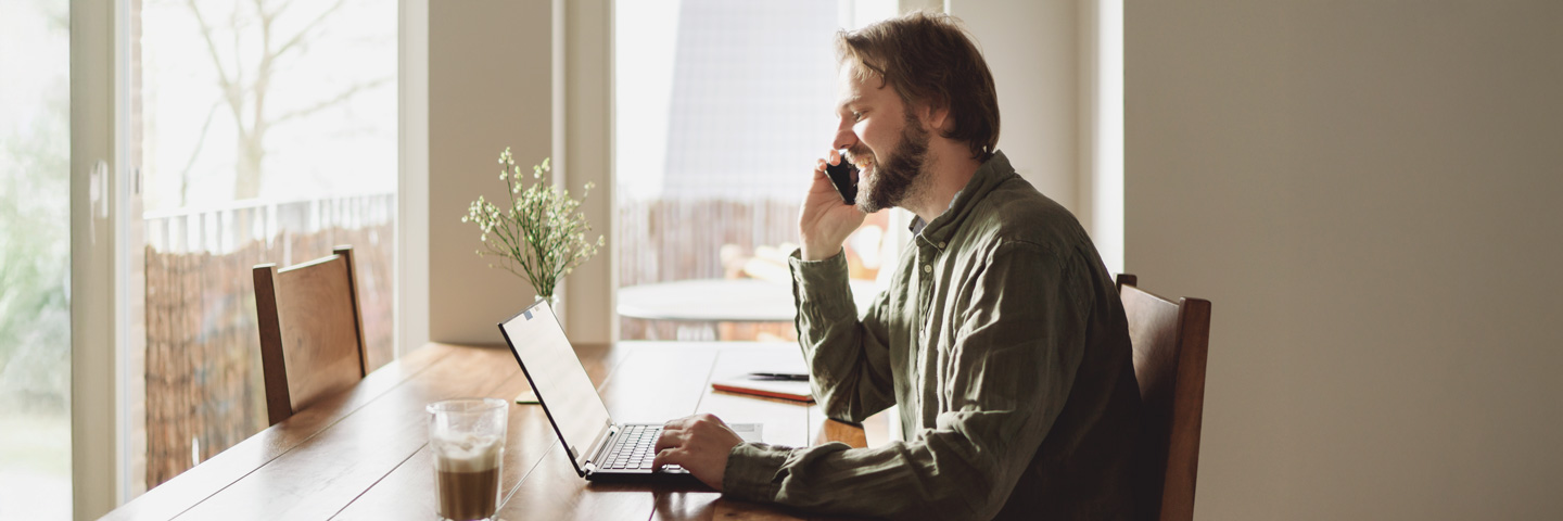 Arbeiten von zuhause: Mann am Esstisch lachend telefoniert und an seinem Notebook arbeitet. Arbeiten von zuhause: Mann am Esstisch lachend telefoniert und an seinem Notebook arbeitet.