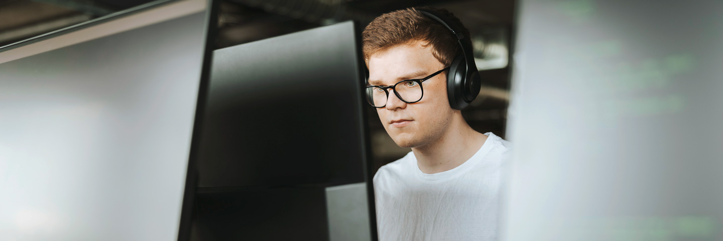 Neurodiversität: Blick auf einen jungen Mann durch Monitore hindurch, der hochkonzentriert ist und Kopfhörer trägt.