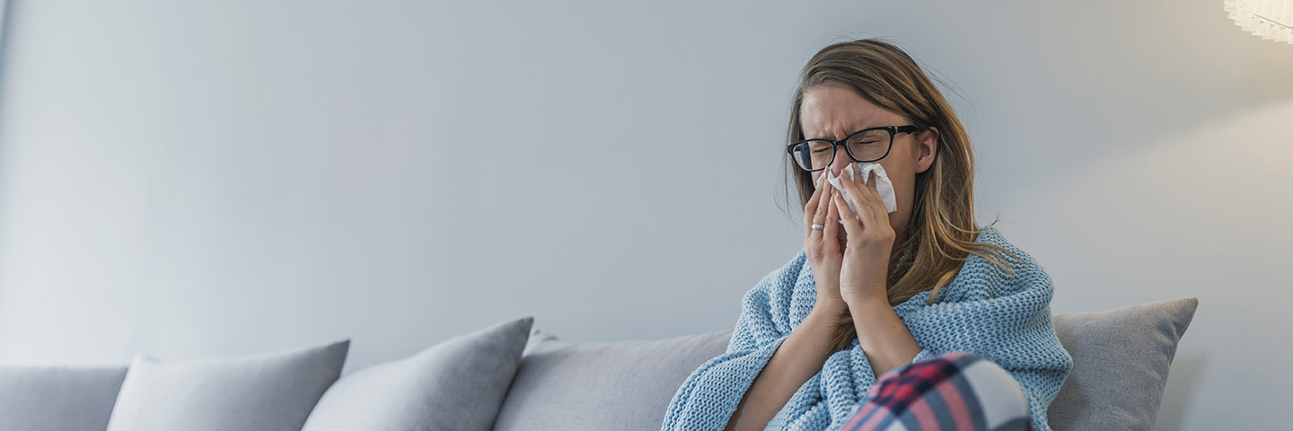 Gesundheitsgerechte Bedingungen im Betrieb helfen Fehlzeiten zu vermeiden Eine kranke Frau sitzt auf einem Sofa und putzt sich die Nase
