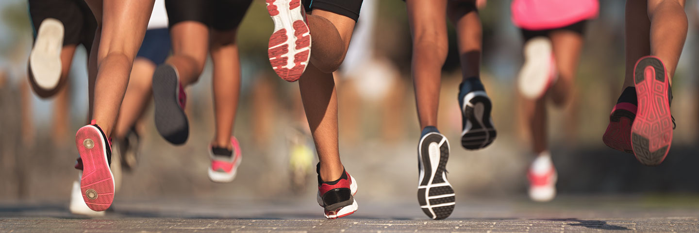 Sport im Betrieb anzubieten, ist ein exzellenter Weg zur Gesunderhaltung der Beschäftigten beizutragen. Die Füße von einer Laufgruppe, die auf einer Straße läuft.